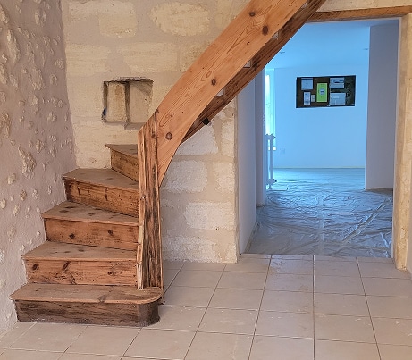 Escalier en bois rustique sous rénovation, près d'un mur en pierre apparent et d'une ouverture vers une pièce lumineuse avec bâche de protection au sol.