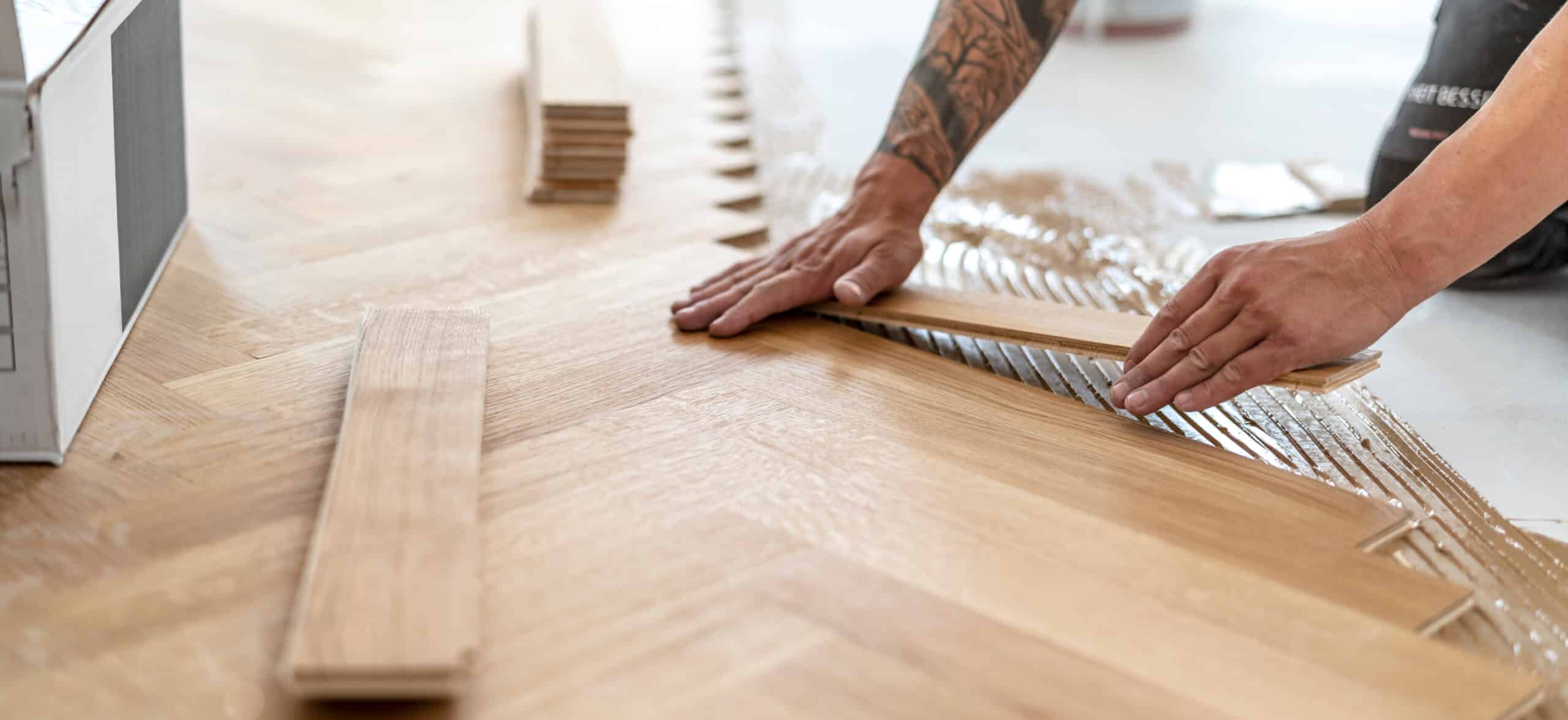Pose de parquet en chevrons en bois clair, un artisan tatoué applique une colle liquide avec ses mains.