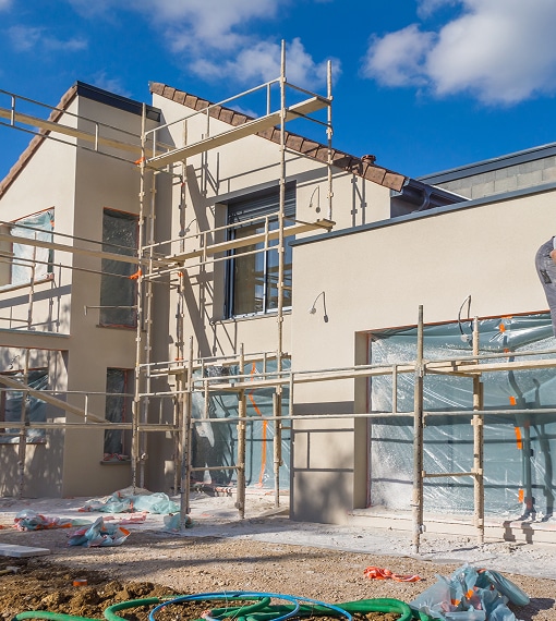 Rénovation façade maison : échafaudage et travaux en cours Chantier de rénovation avec échafaudage monté devant une façade beige, fenêtres bâchées sous un ciel bleu et nuageux.